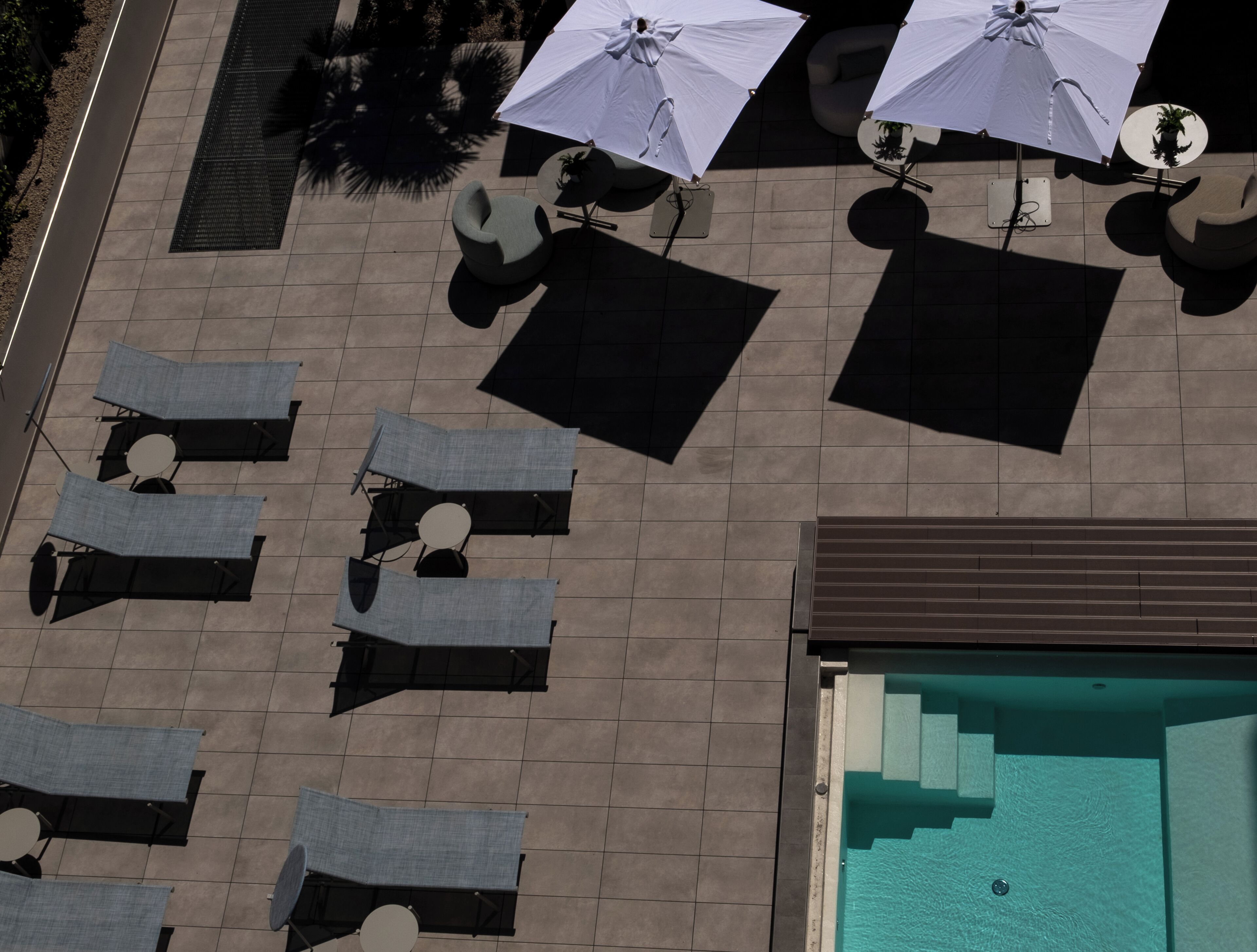 An aerial view of a rooftop pool area with multiple lounge chairs and umbrellas, a swimming pool, and a tiled deck.