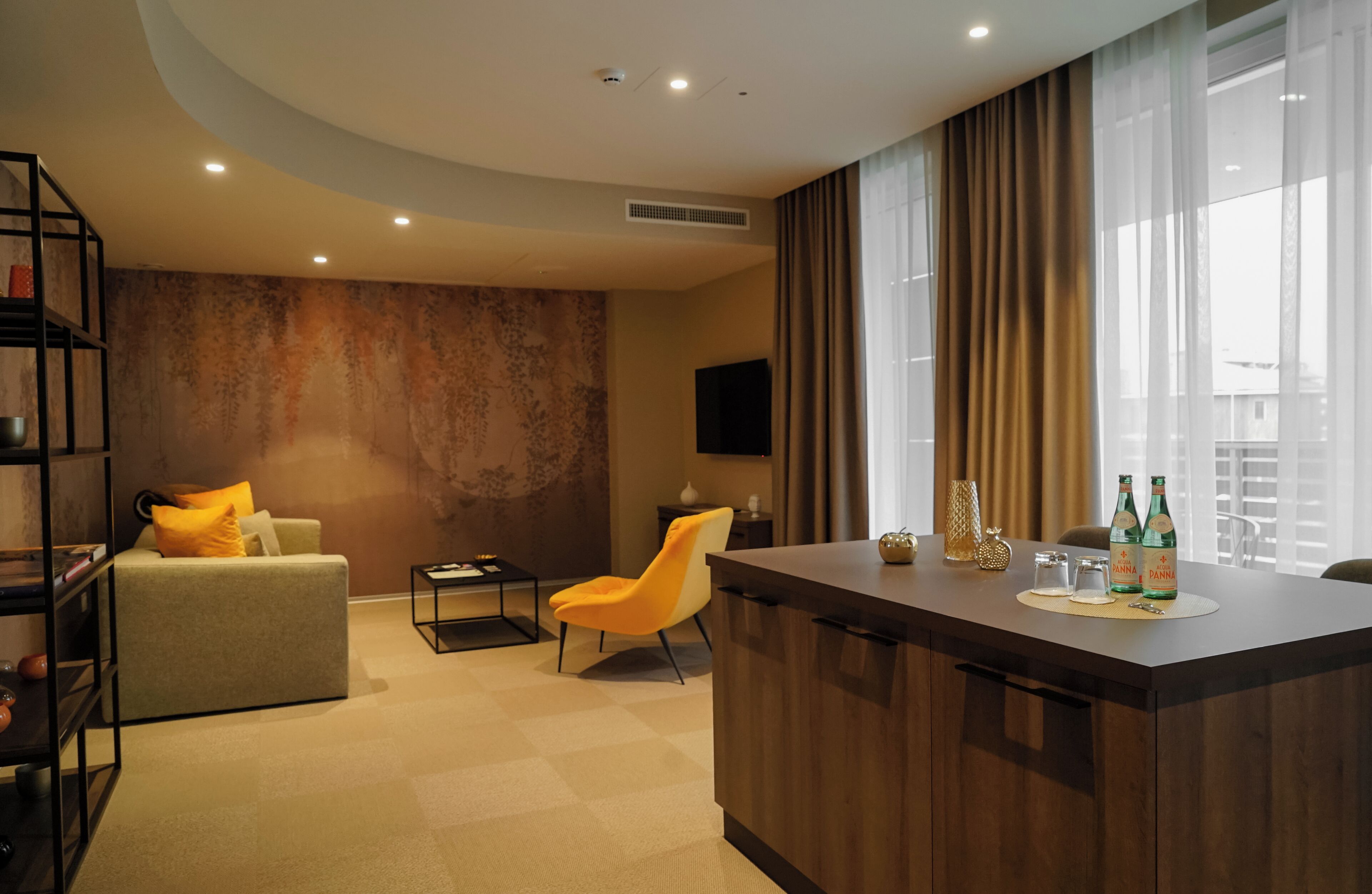 A modern hotel room with a sofa, a chair, a small table, a bar area, and a TV mounted on the wall.