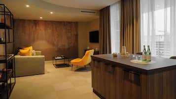 Una habitación de hotel moderna con sofá, una silla, una mesita, una zona de bar y un televisor montado en la pared.