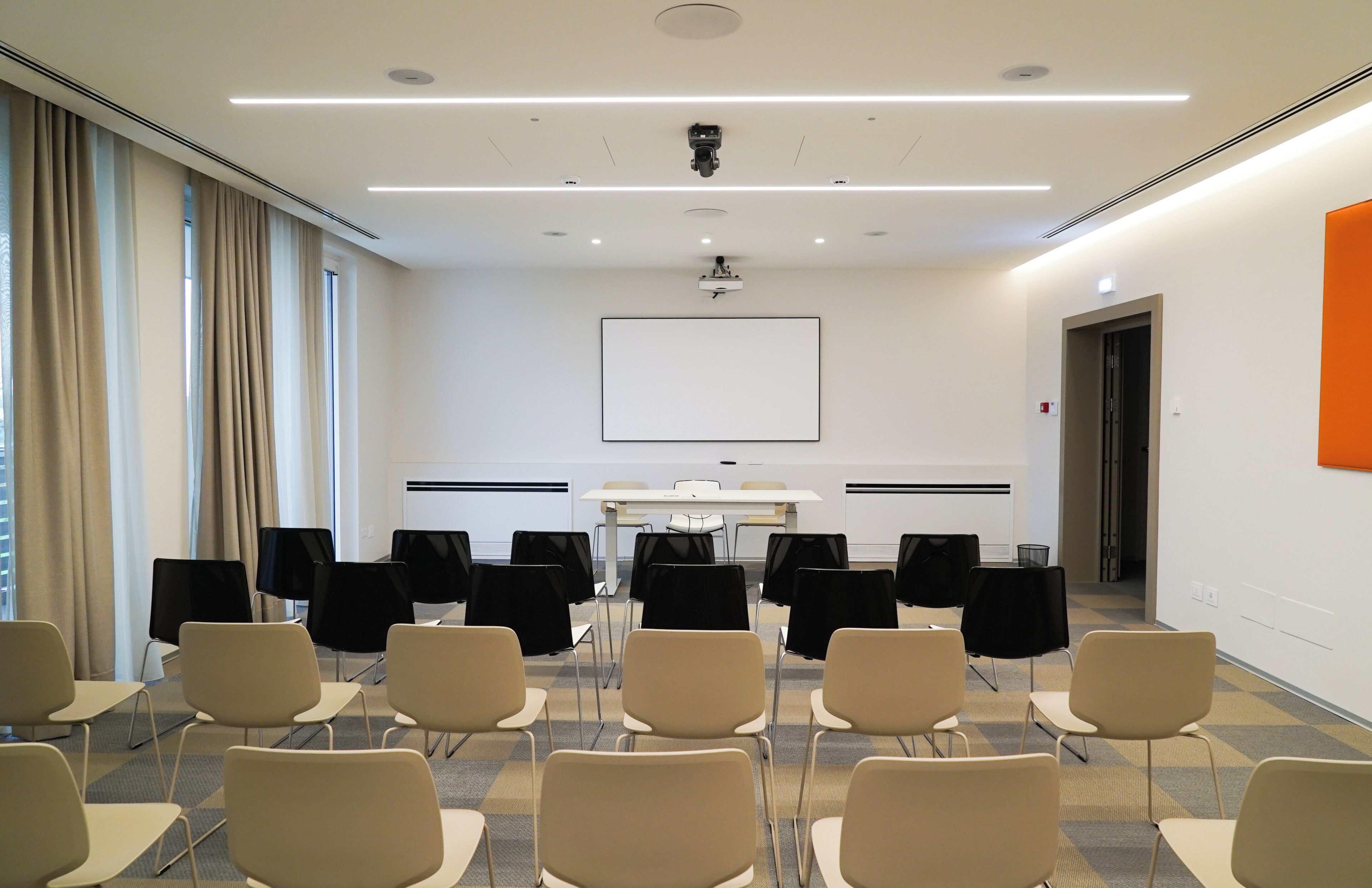 A conference room with a white table, black chairs, a projector screen, and a large window with curtains.