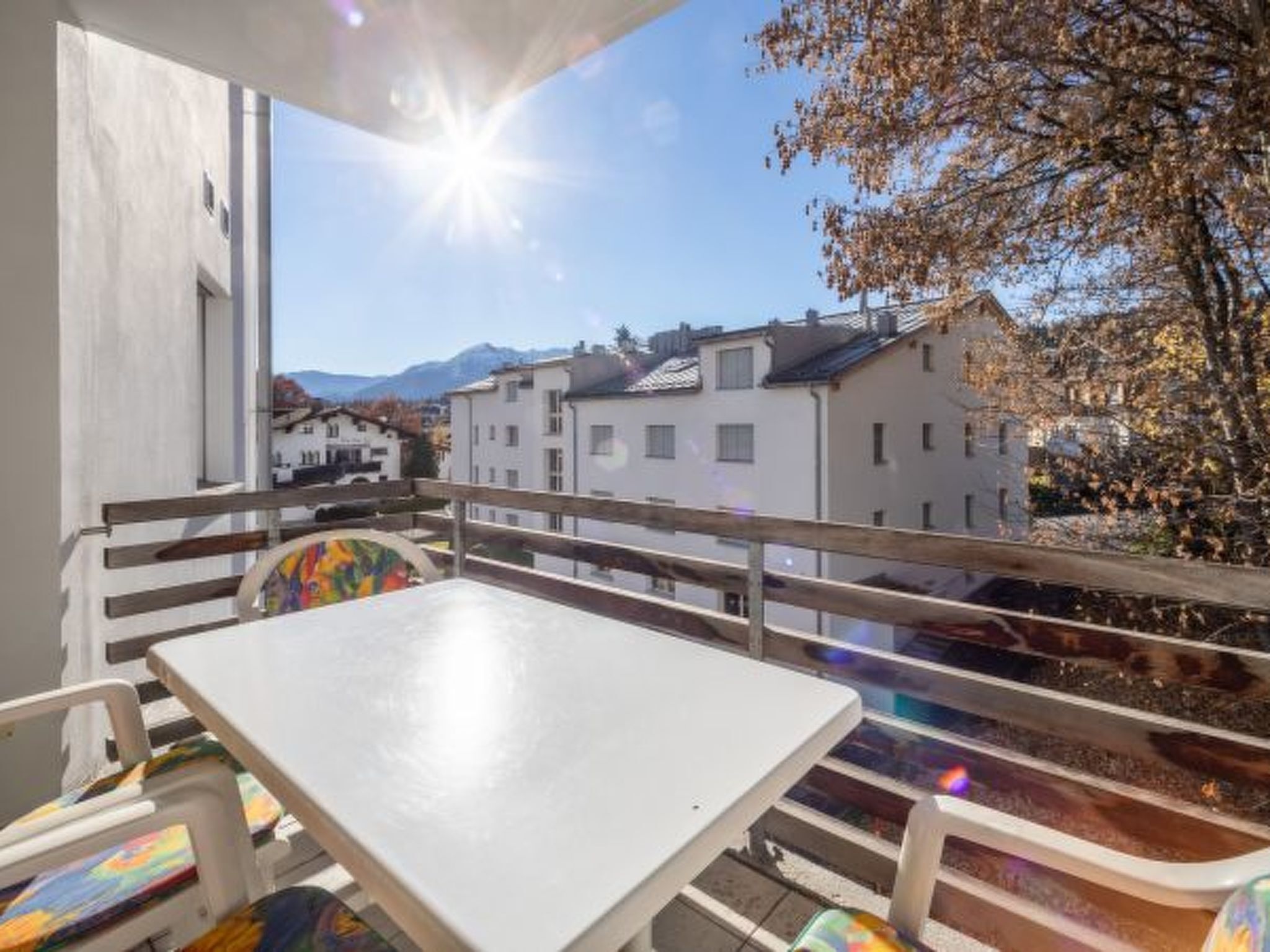 A balcony with a white table and chairs, overlooking a residential area with houses and mountains.