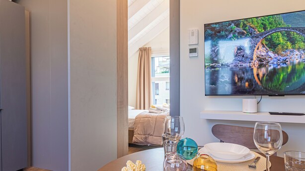A hotel room with a bed, a TV mounted on the wall, and a dining table set with glasses and plates. - Flower Residence - Appartamento Iris (Lissone)