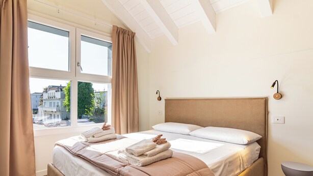 A bedroom with a bed, window, curtains, bedside table, and a view of buildings outside. - Flower Residence - Appartamento Lavanda (Lissone)