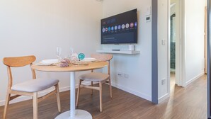 A modern dining area with a round table, two wooden chairs, a flat-screen TV mounted on the wall, and a hallway leading to another room. - Flower Residence - Appartamento Lavanda (Lissone)