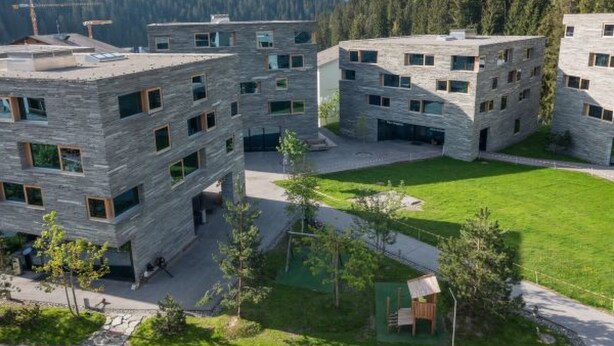 Een modern gebouw met meerdere verdiepingen en grote ramen, omgeven door een verzorgd gazon en bomen, met bergen op de achtergrond.