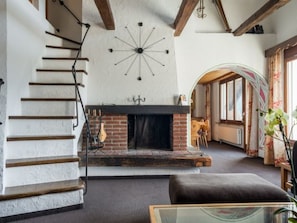 Een trap met zichtbare houten balken, een openhaard met bakstenen omlijsting, en een grote, gewelfde doorgang naar een andere kamer.