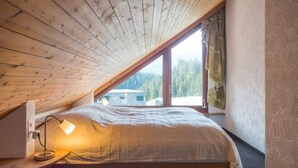 Een slaapkamer met een schuine kap, een bed, een bedlamp en een raam met uitzicht op een huis en bomen.