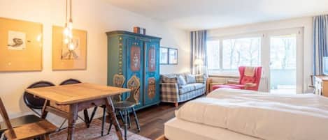 Una habitación con una cama, una mesa de madera con sillas, un armario azul, una silla roja y una ventana con cortinas.