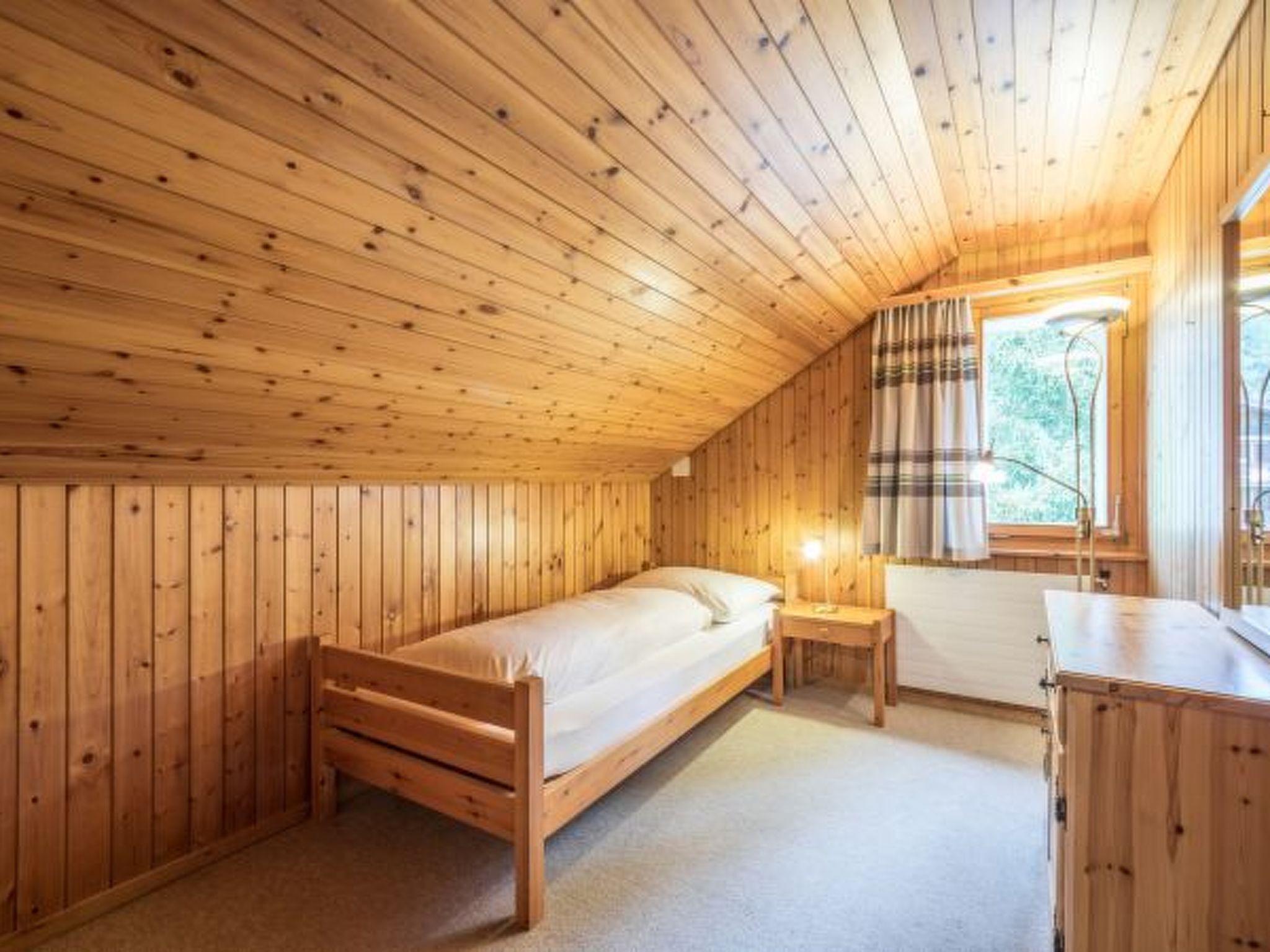 A wooden cabin room with a single bed, a nightstand, and a window with curtains.