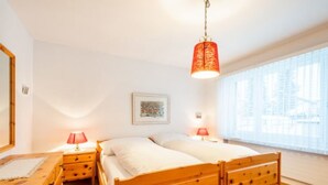A bedroom with two wooden beds, bedside lamps, a bedside table, a large window with a view, and a hanging light fixture. - Via Sulten 12 by Interhome (Flims)