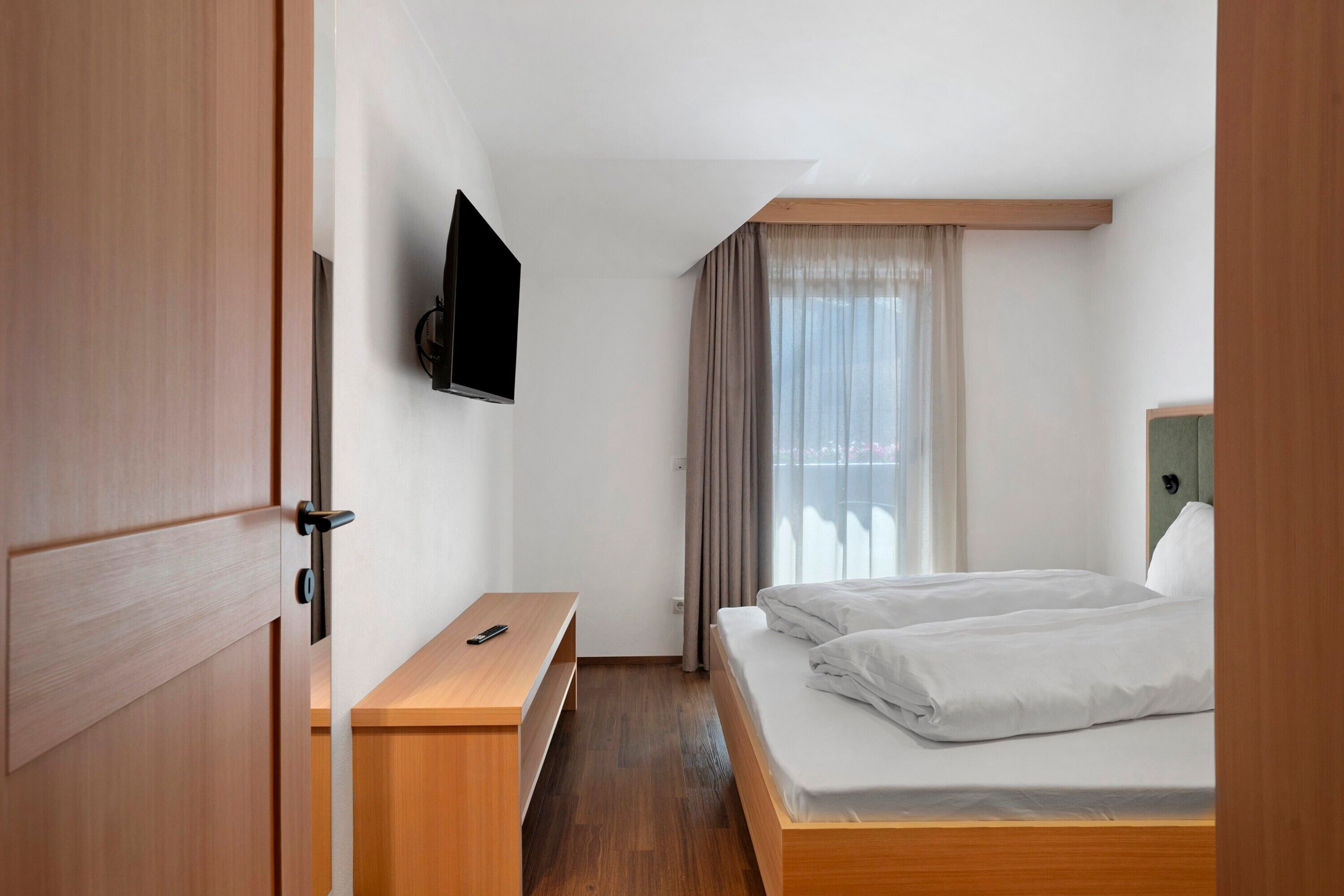 Quarto de hotel com uma cama, televisão fixada na parede, mesa de cabeceira e uma janela com cortinas.