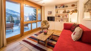 Una sala acogedora con un sofá rojo, una mesa de centro de madera y un sillón. Se observa una estantería con diversos objetos y un amplio ventanal con vistas a una montaña nevada.