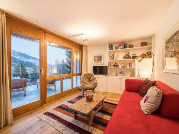 A cozy living room with a red sofa, a wooden coffee table, and a chair. There is a bookshelf with various items and a large window offering a view of a snowy mountain.