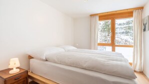 A bedroom with a bed, a nightstand, a window with curtains, and a view of snow outside. - Casa Collina 5i-02 by Interhome (Flims)