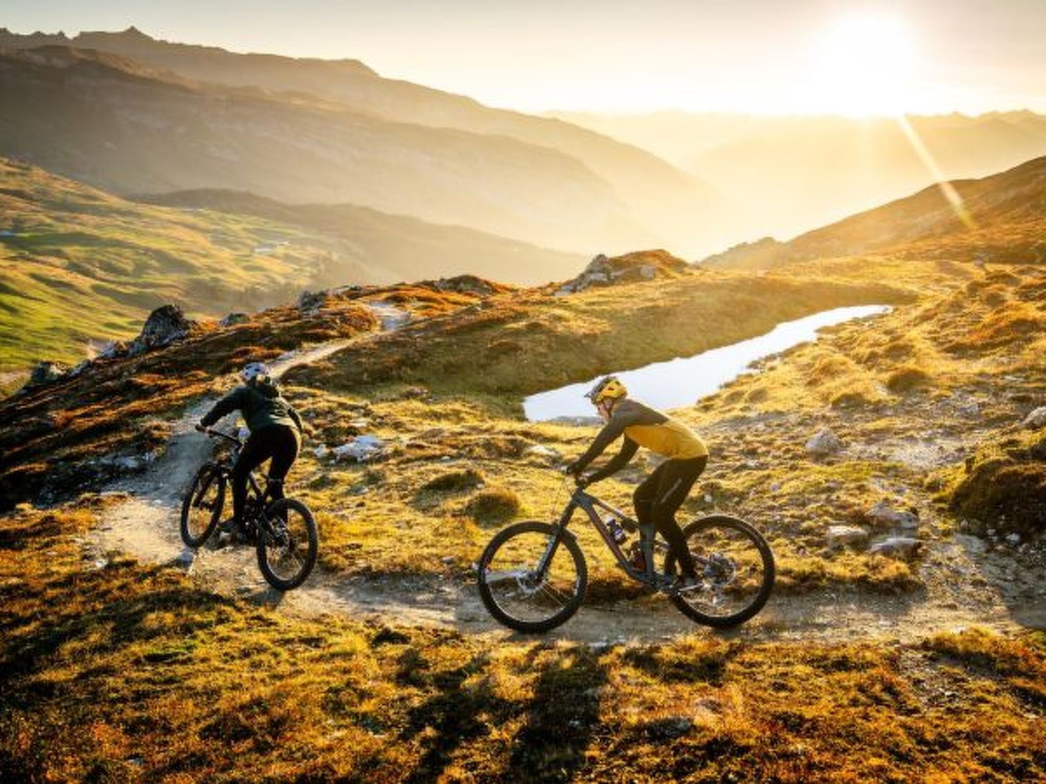 Deux cyclistes sur un sentier de montagne, sous un ciel dégagé, avec des montagnes qui se dessinent à l’horizon.