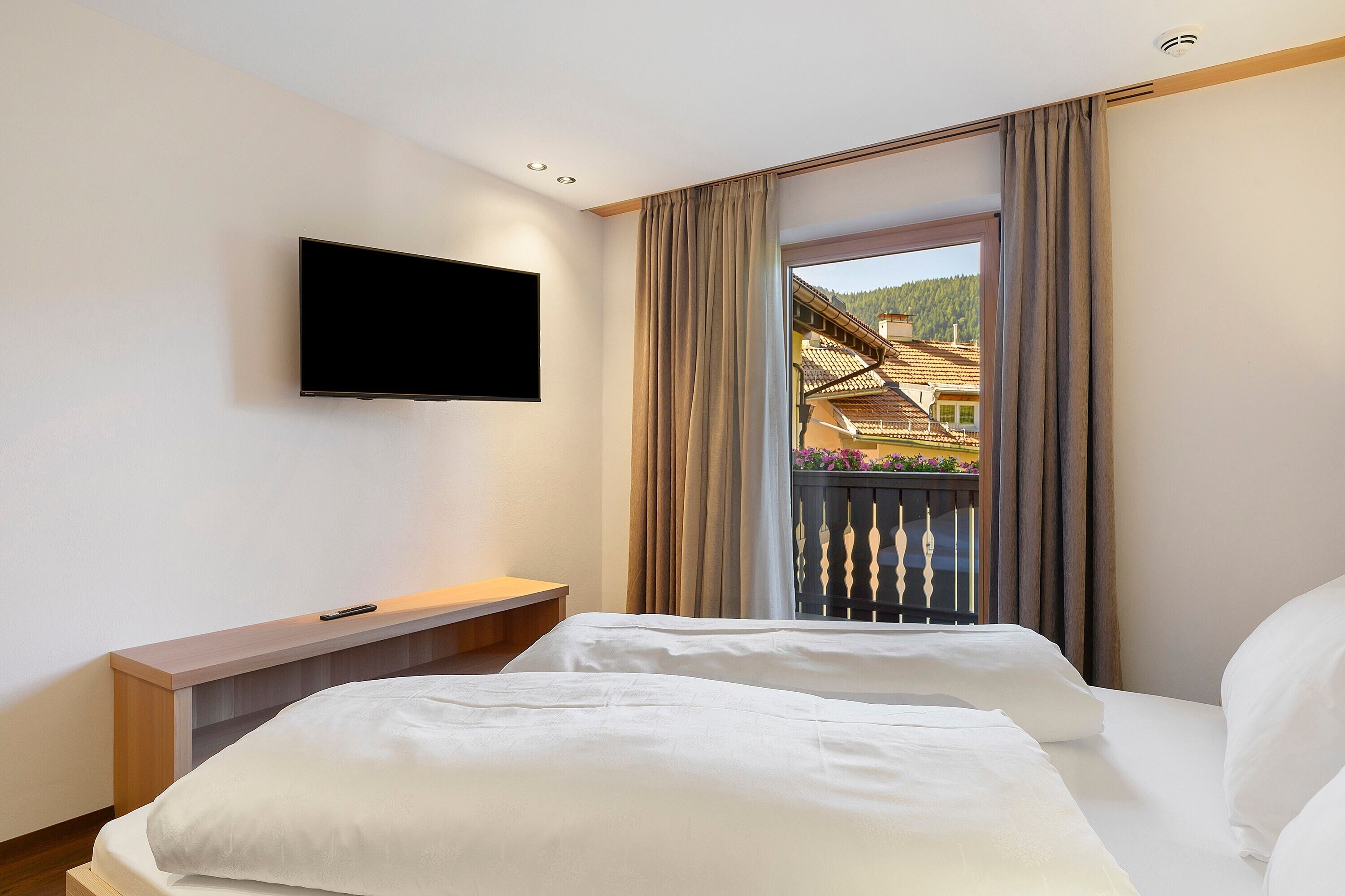 A hotel room with a bed, a TV mounted on the wall, and a balcony with a view of buildings.