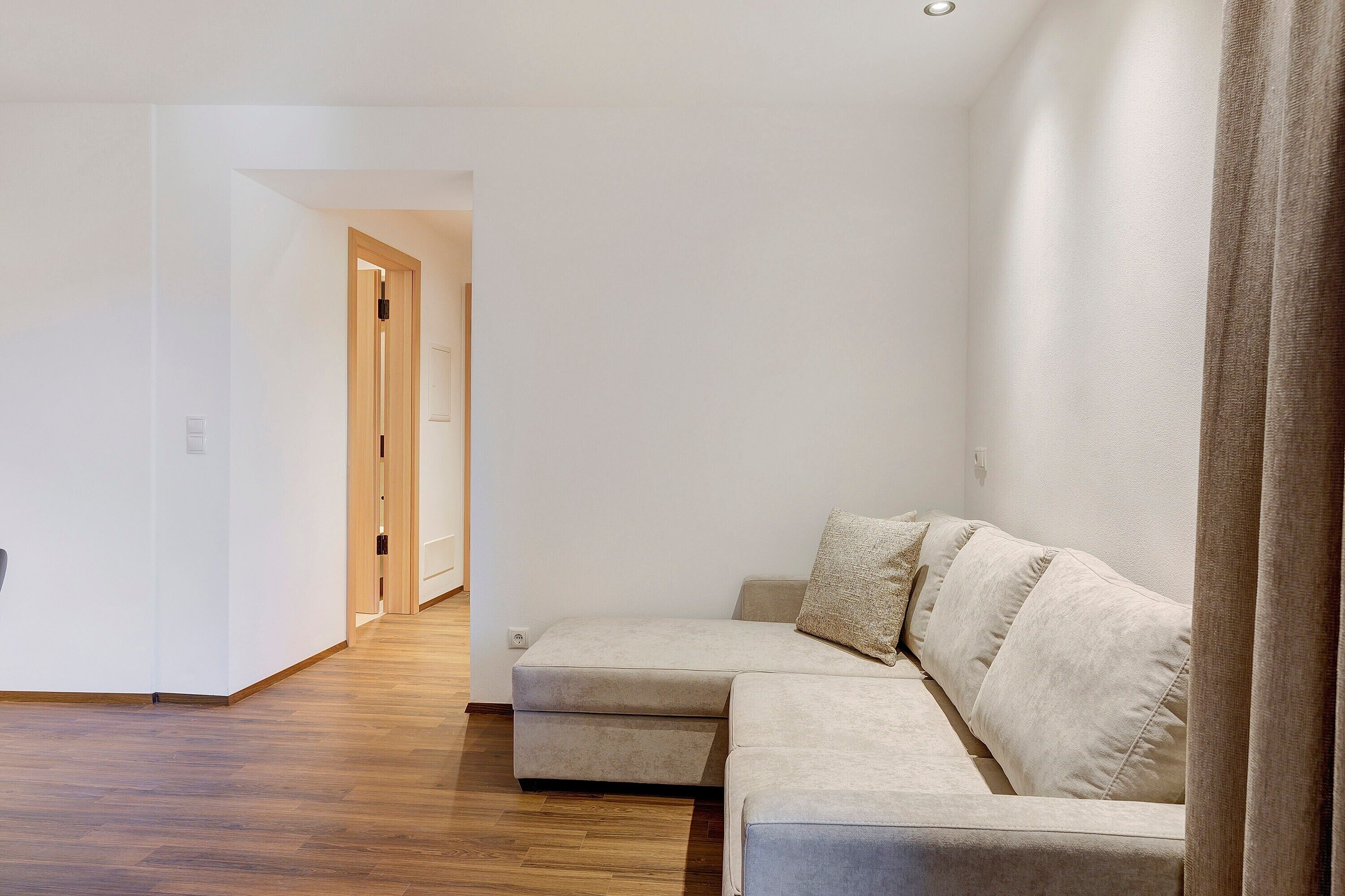 Un salon moderne avec un canapé d’angle, un plancher en bois et une porte donnant sur une autre pièce.