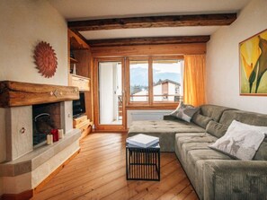 Ein Wohnzimmer mit Kamin, Holzbalken, einer Couch und Blick auf Gebäude durch große Fenster.