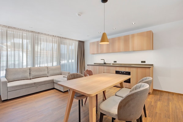 Un salon moderne comprenant un canap é, une table à manger, des chaises et un coin cuisine avec des meubles en bois et un luminaire suspendu.