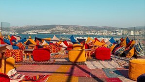 Een terras op het dak met kleurrijke kussens en vloerkleden, met uitzicht op een stadsbeeld en heuvels.