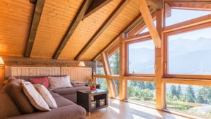 Accogliente baita con soffitto in legno, un divano, un tavolino e ampie finestre che si affacciano su una vista montana.