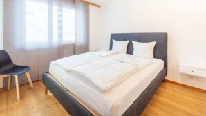 Ein Schlafzimmer mit einem Bett, einem Sessel, einem Fenster mit Vorhängen und einem Holzboden.