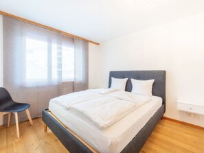 A bedroom with a bed, a chair, a window with curtains, and a wooden floor. - Casa Guarda Val 1 by Interhome (Falera)