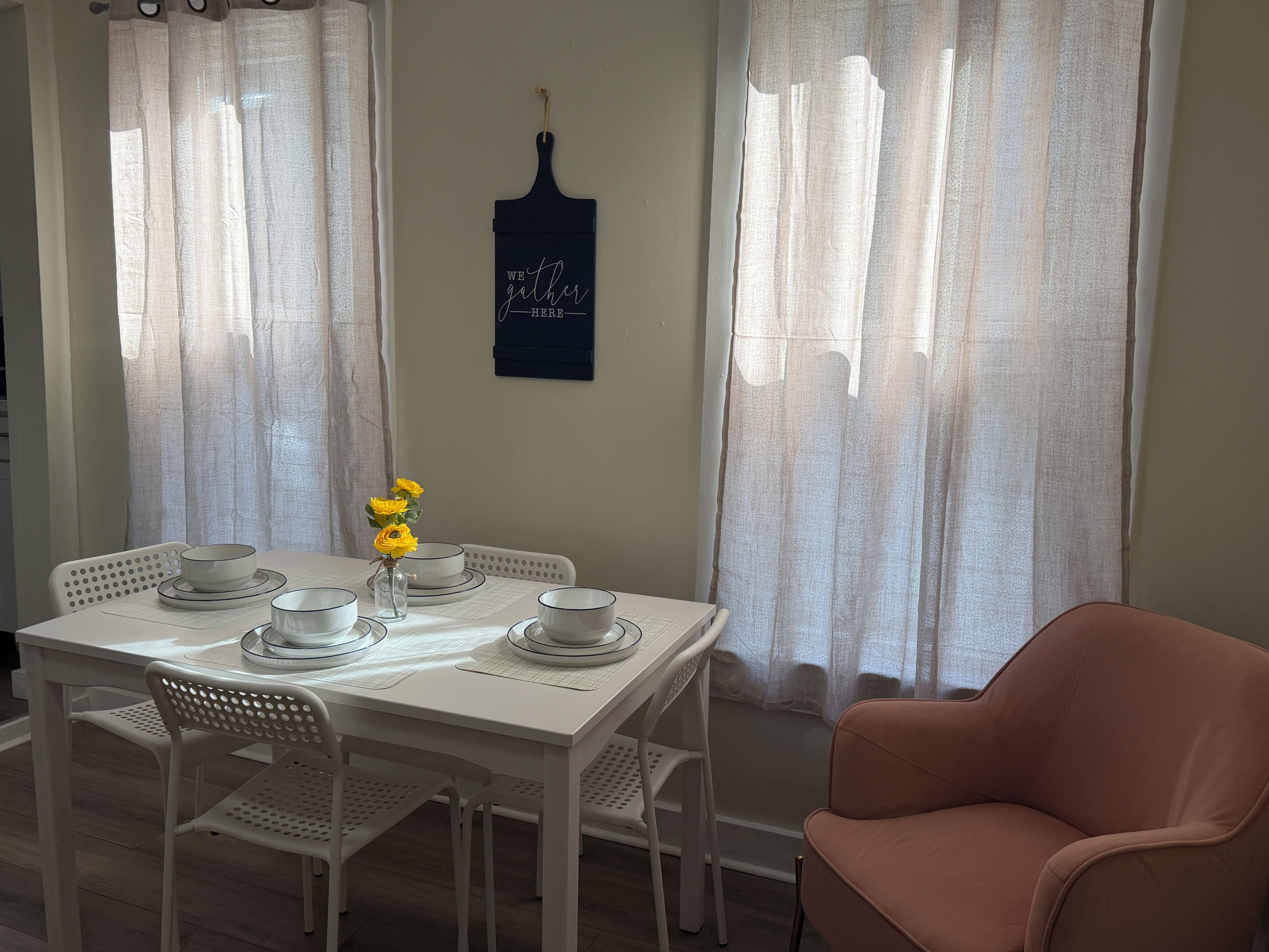 Un coin repas avec une table blanche dressée pour quatre personnes, un vase rempli de fleurs jaunes et un panneau bleu foncé accroché au mur.