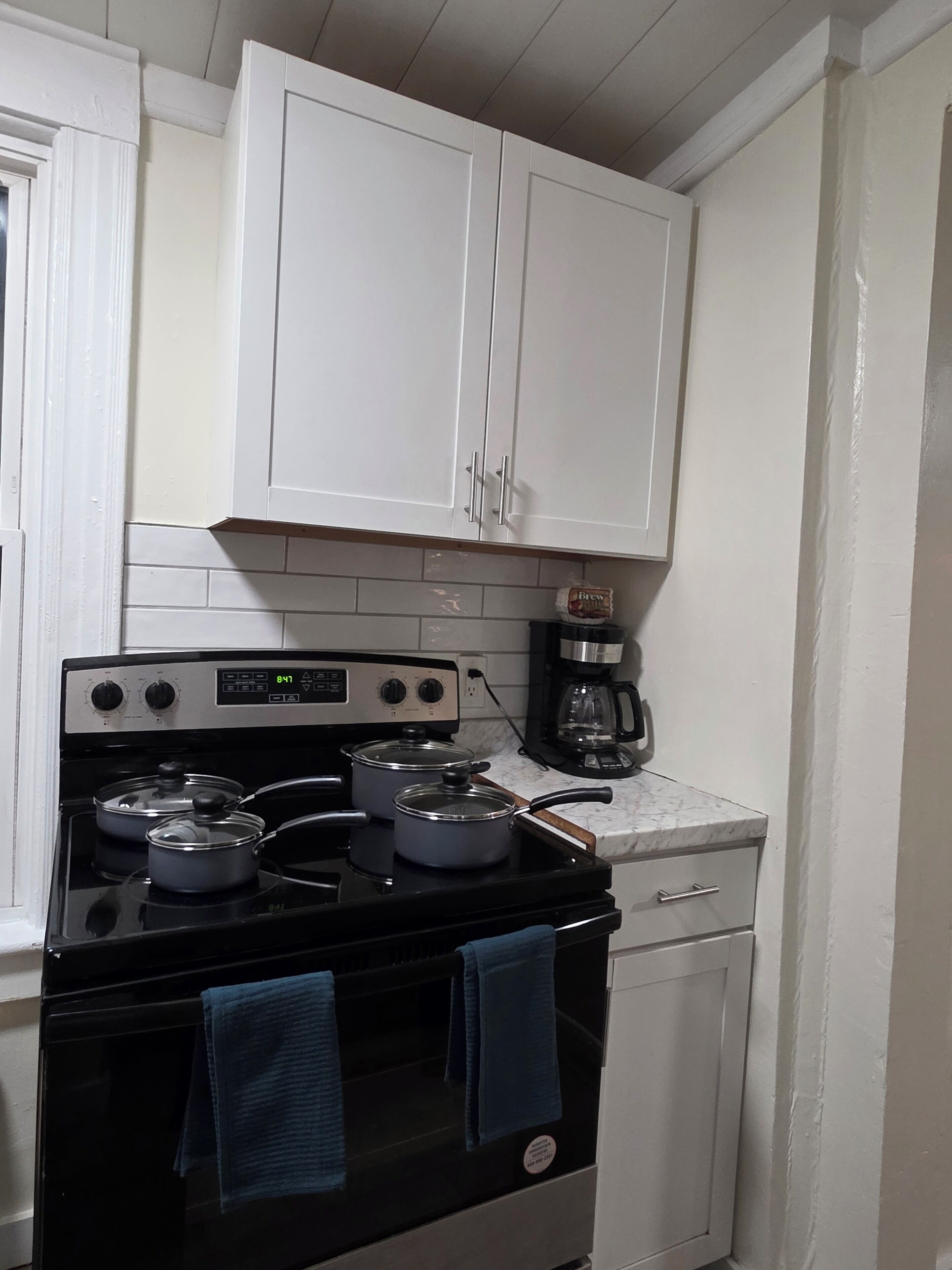 Une cuisine avec des meubles blancs, une cuisinière avec des casseroles, une machine à café et une fenêtre.