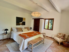 Une chambre à coucher avec un lit, un banc, une chaise, une lampe, un tableau et une fenêtre.