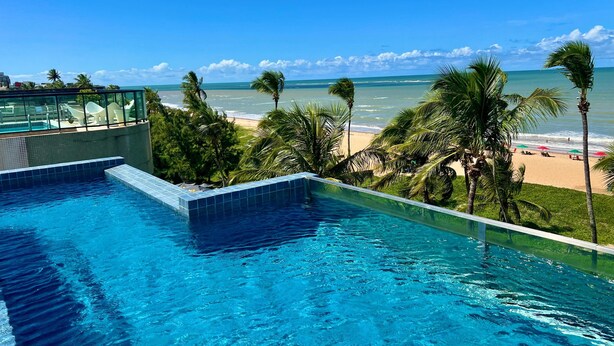 Ein Schwimmbecken mit klarem, blauem Wasser, ein Strand mit Palmen und Blick auf den Ozean.