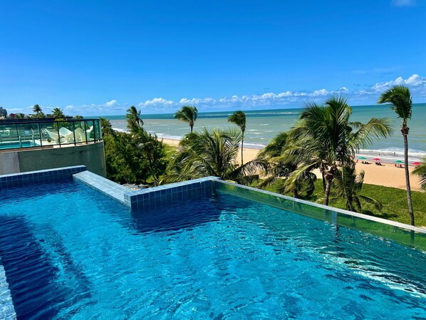 Ein Schwimmbecken mit klarem, blauem Wasser, ein Strand mit Palmen und Blick auf den Ozean.