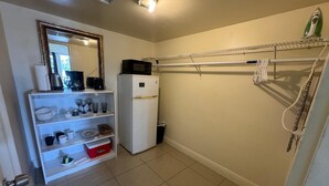 Cocina compacta con refrigerador blanco, microondas y cafetera sobre la encimera. Hay una repisa con cristalería y una caja roja de almacenamiento. Un tendedero está fijado a la pared.
