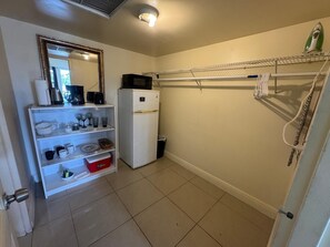 Eine kompakte Küche mit einem weißen Kühlschrank, einer Mikrowelle und einer Kaffeemaschine auf der Arbeitsfläche. Ein Regal mit Gläsern und einer roten Aufbewahrungsbox. Ein Wäscheständer ist an der Wand befestigt.
