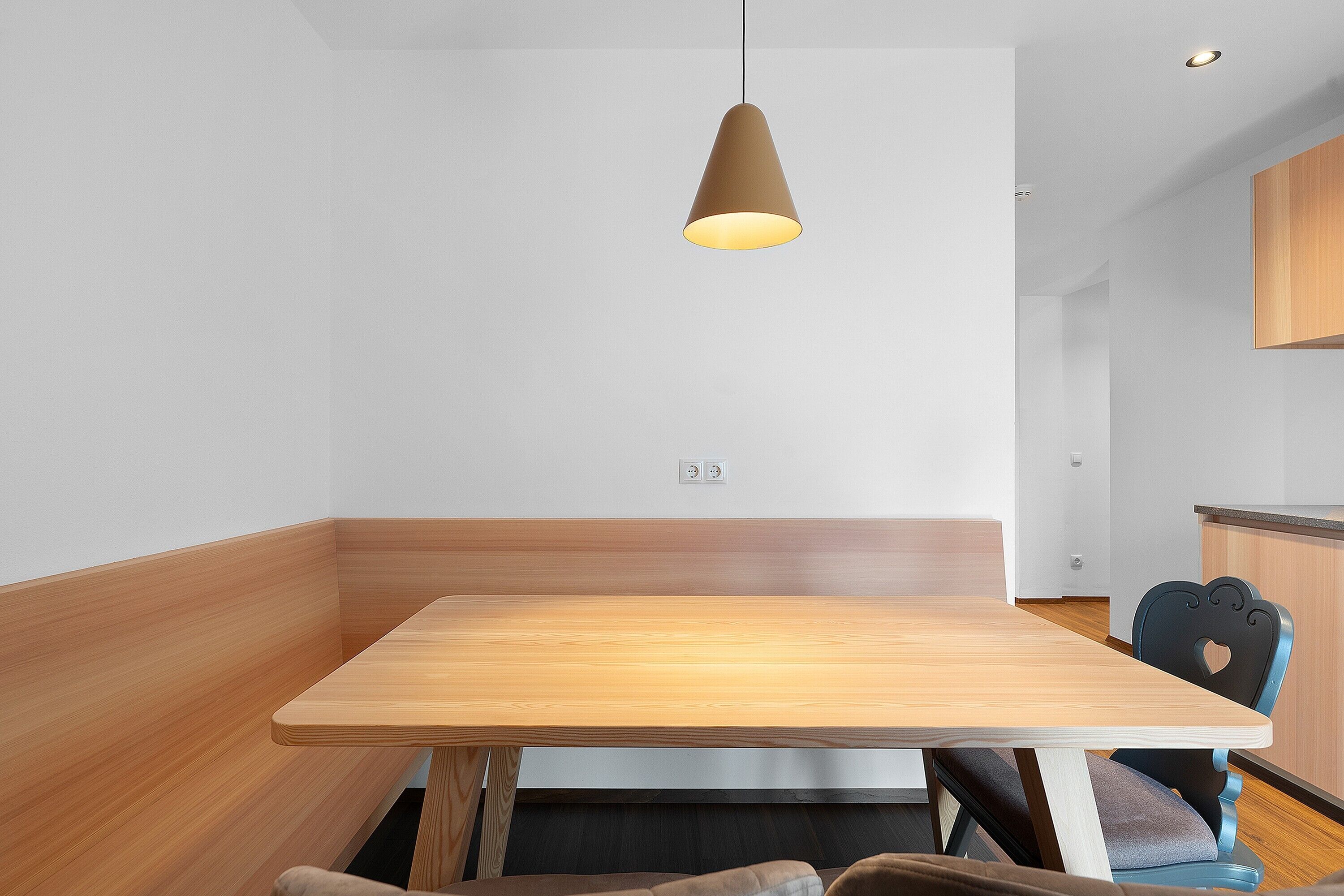 A modern dining area with a wooden table and chairs, a hanging light fixture, and a kitchen area in the background.