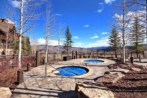 A pool area with two circular pools, surrounded by a stone deck and wooden fencing, with mountains in the background. - NEW! 3BR Condo in Bachelor Gulch, Ski in/out (Avon)