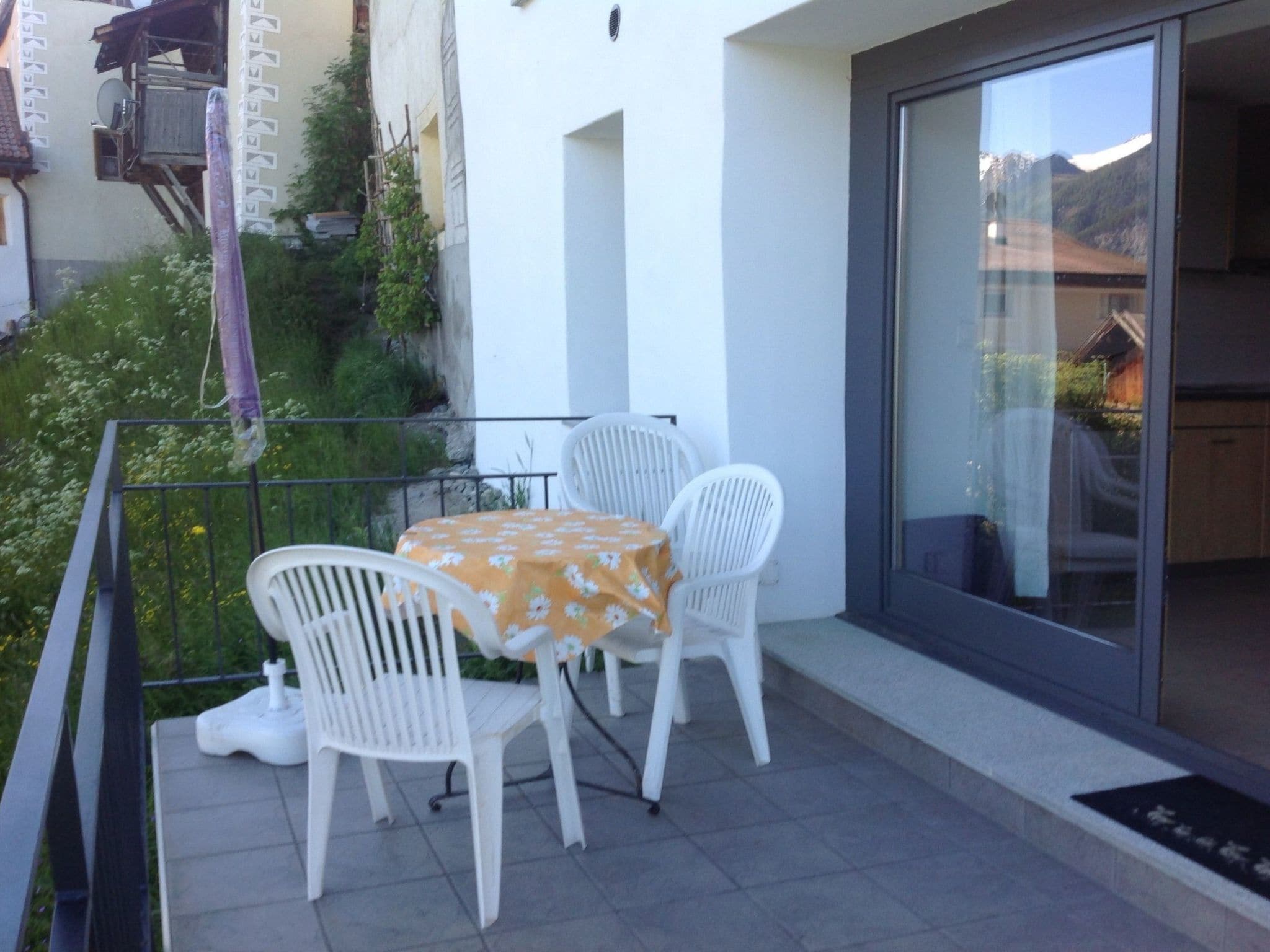 Un balcon avec une table et des chaises, une porte vitrée donnant sur une vue sur les montagnes et un mur blanc orné d’une antenne parabolique.