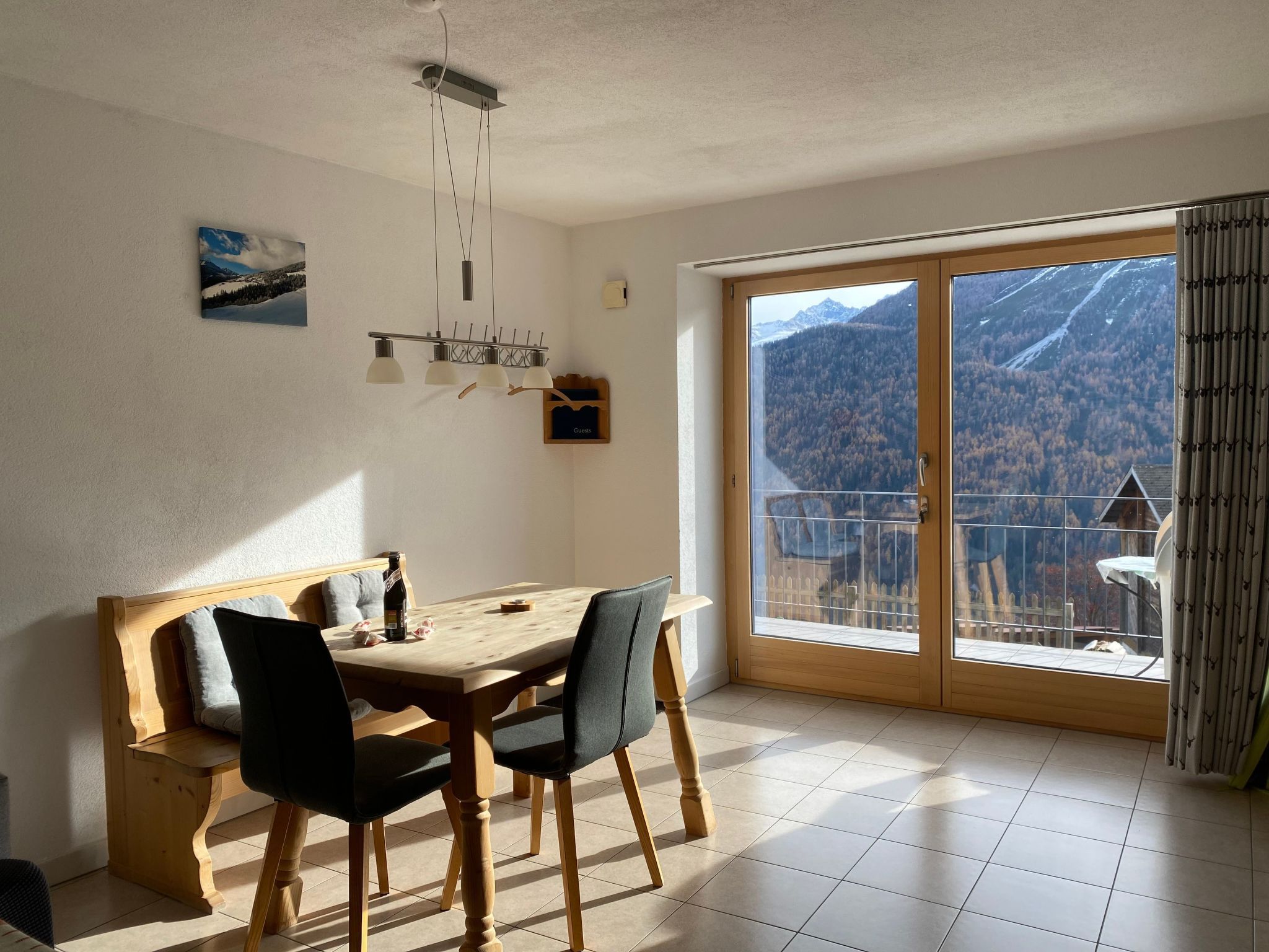 A dining area with a wooden table and chairs, a painting on the wall, and a view of mountains through a glass door.