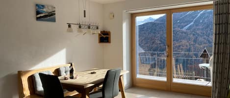A dining area with a wooden table and chairs, a painting on the wall, and a view of mountains through a glass door.