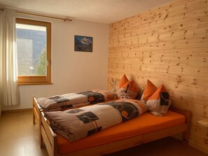 Une chambre avec une tête de lit en bois, deux lits avec une literie orange, une fenêtre donnant sur des arbres et un tableau accroché au mur.