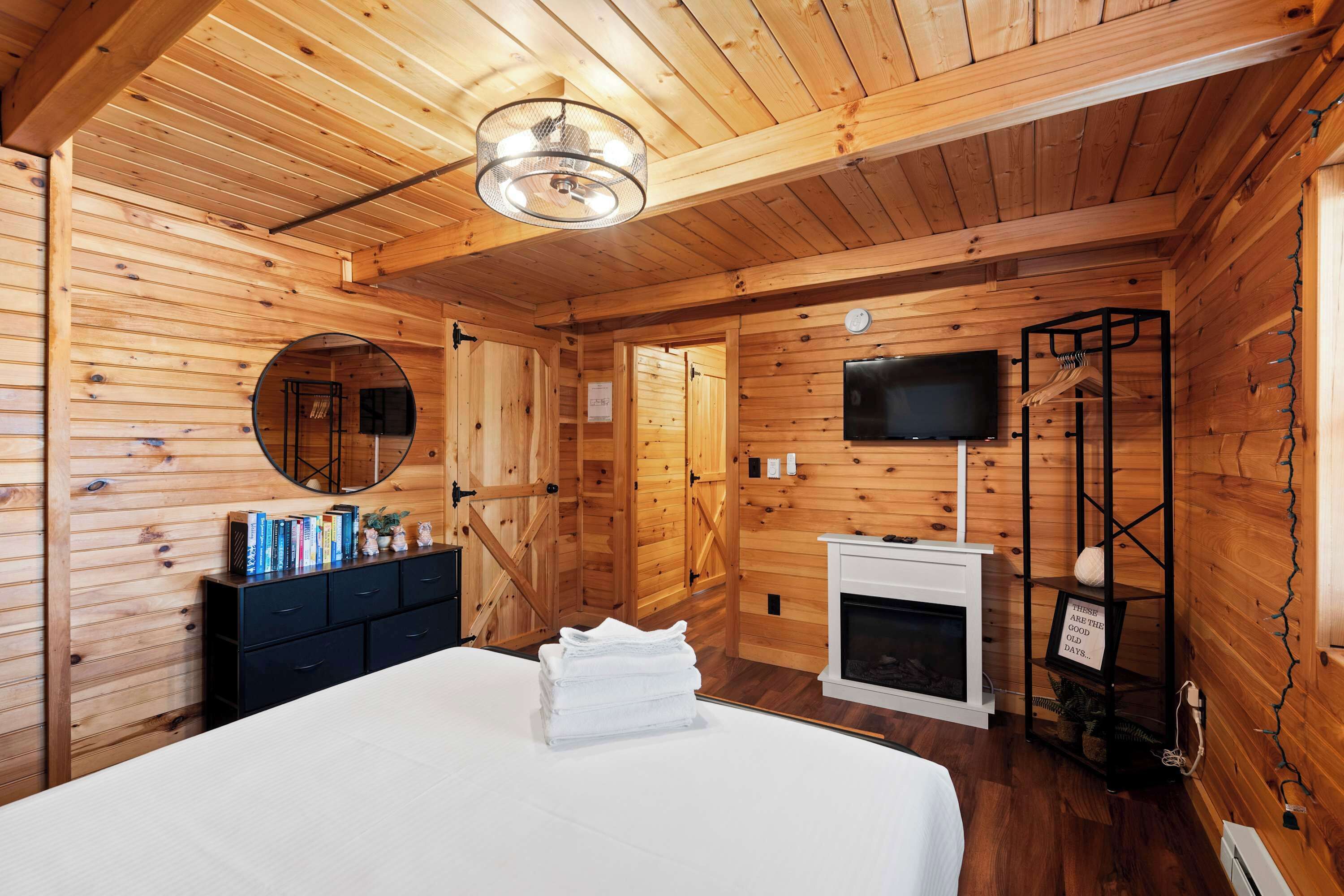 L’intérieur d’un chalet en bois comprend un lit, une commode, un miroir, une cheminée, un téléviseur et une étagère sur laquelle est posé un panneau.