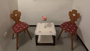 Ein Zimmer mit einem Holzkabinett, zwei Stühlen mit floralem Bezug, einem kleinen Tisch mit einer Vase und einer weißen Wand.