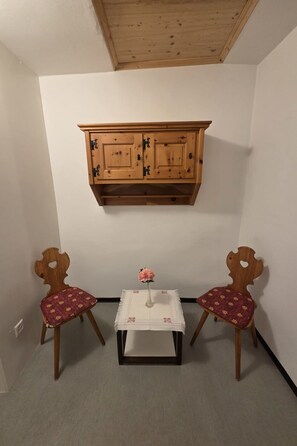 Ein Zimmer mit einem Holzkabinett, zwei Stühlen mit floralem Bezug, einem kleinen Tisch mit einer Vase und einer weißen Wand.