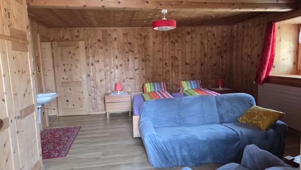 Ein Zimmer mit Holzboden, einem Bett, einem Schlafsofa, einem Nachttisch, einer Lampe und einem Fenster mit Vorhängen.