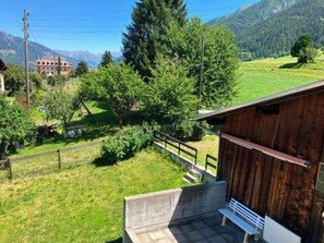 Una cabaña de madera con un banco y una plataforma de concreto, rodeada de un área verde con árboles y una montaña al fondo.