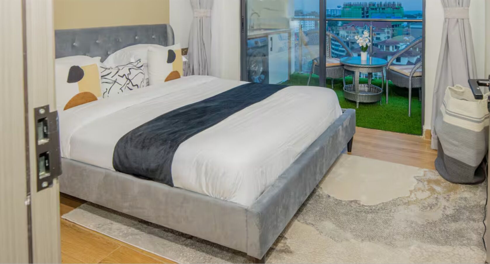 A bedroom with a bed, a view of the city, a balcony with a table and chairs, and a trash bin.