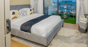 Ein Schlafzimmer mit Bett, Blick auf die Stadt, einem Balkon mit Tisch und Stühlen sowie einem Mülleimer.