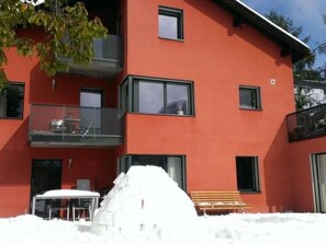 Ein mehrstöckiges rotes Gebäude mit Balkonen, einem verschneiten Untergrund und einer Bank.