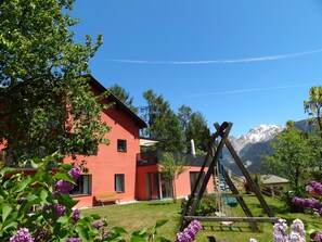 A red building with a wooden swing set in a garden, surrounded by mountains and trees. - Chasa Felix, (Scuol) by Interhome (Scuol)