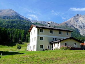 A two-story building with a sloped roof, surrounded by a grassy field and mountains in the background. - Villa Kuntner Bund, (Tschierv) by Interhome (Tschierv)